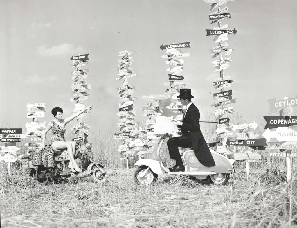 1960 man and woman with two Lambretta scooters in front of a lot of signs with cities